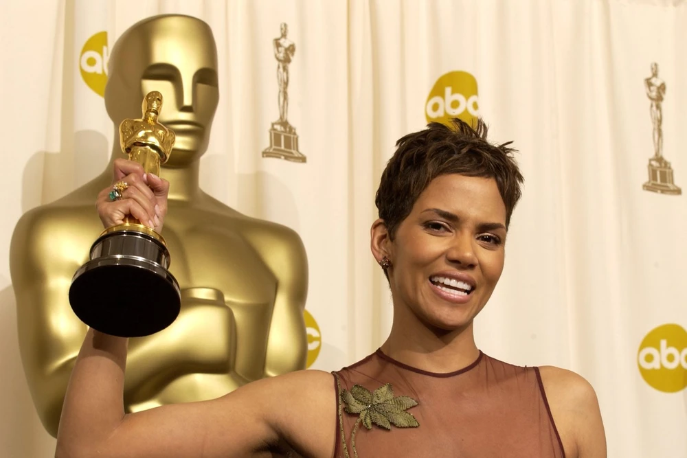 Лауреат премии Оскар держит статуэтку на фоне золотой статуи и логотипов ABC, улыбаясь в элегантном платье.