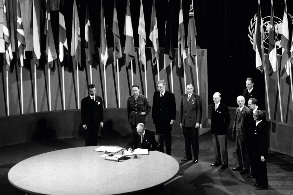 Исторический момент: подписание Устава ООН. Делегат за круглым столом подписывает документ в присутствии других представителей стран. На заднем плане — множество национальных флагов.