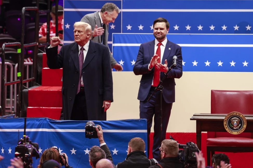 Два политика на сцене с американским фоном со звёздами. Один поднимает кулак, другой аплодирует у микрофона. Видна президентская печать США.