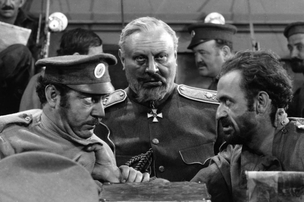 Чёрно-белый кадр из фильма: мужчина в военной форме с крестом на груди в центре, рядом двое солдат, на фоне другие люди.