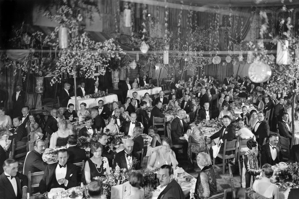Чёрно-белое фото официального банкета 1920-30-х годов: гости в смокингах и вечерних платьях за столами в богато украшенном зале.