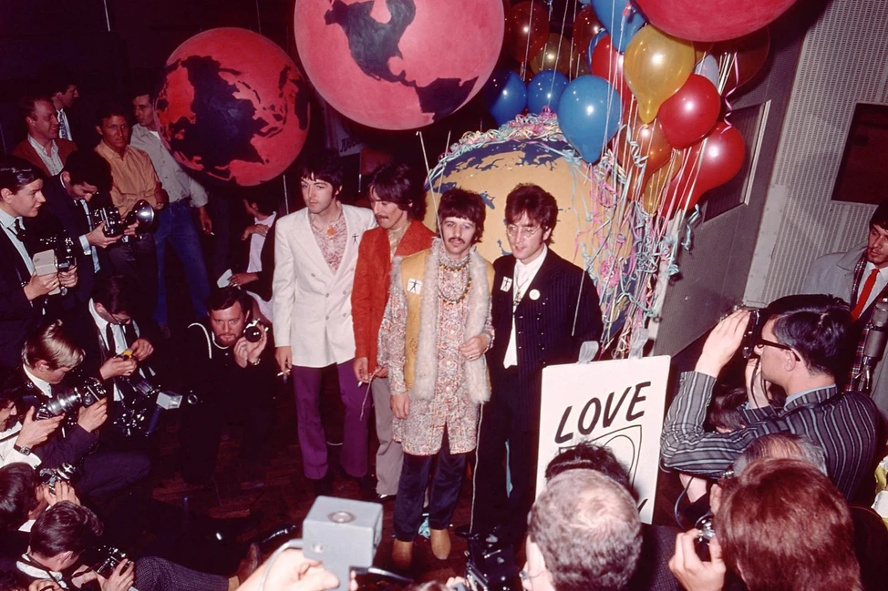 The Beatles в ярких костюмах среди шаров-глобусов и знака «LOVE» в окружении фотографов.