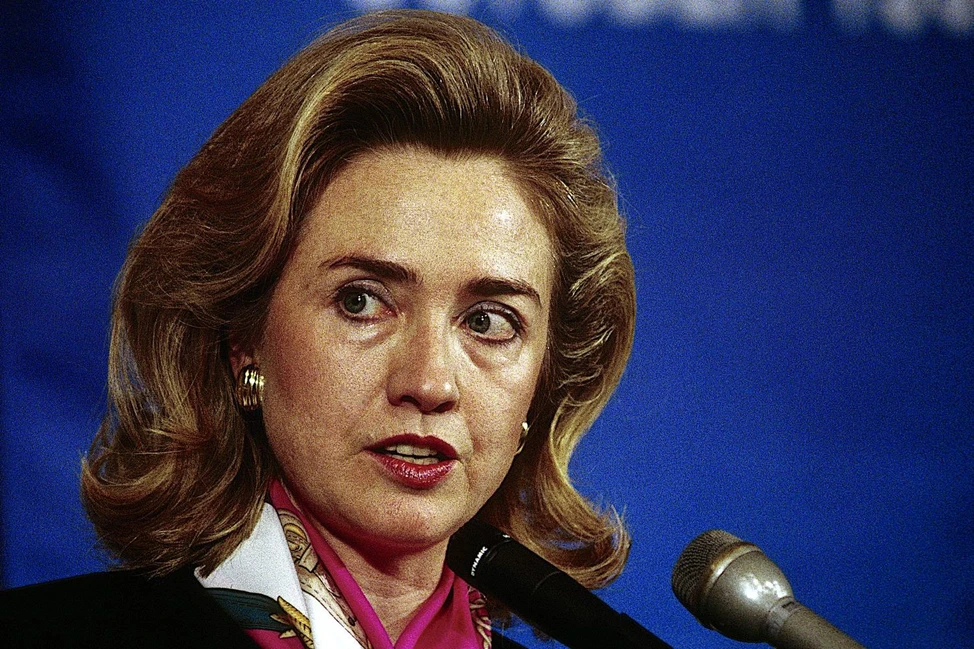 Хиллари Клинтон выступает у микрофона на синем фоне. Крупный план женщины в тёмном пиджаке и розовом шарфе.