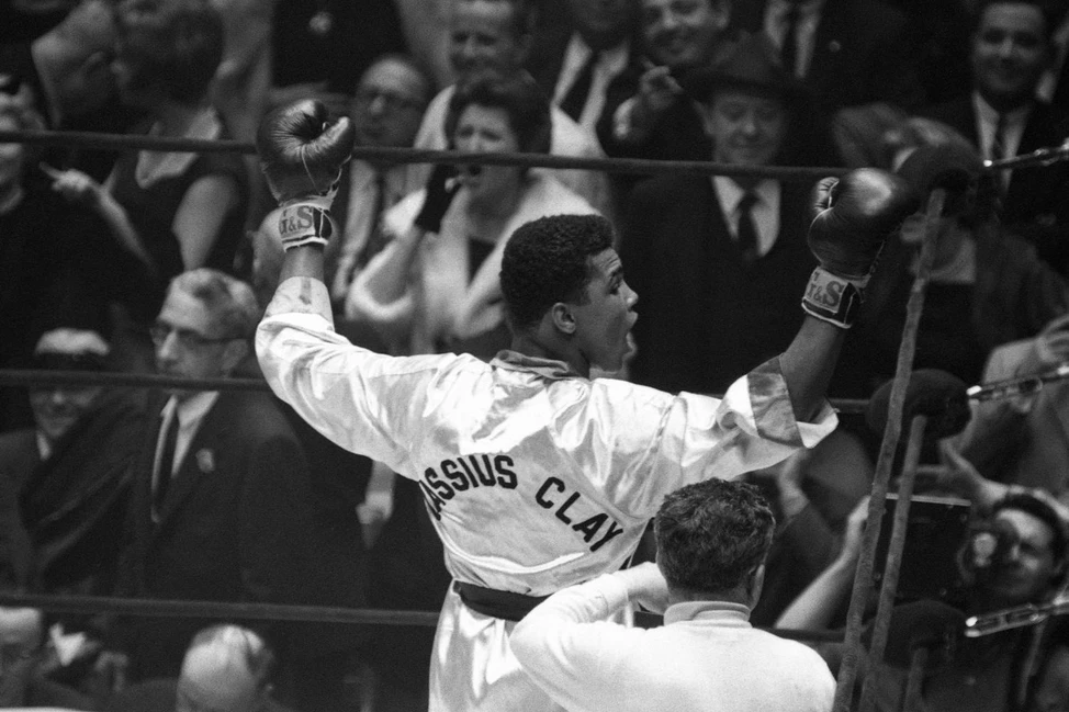 Кассиус Клей (будущий Мухаммед Али) празднует победу на боксёрском ринге, подняв руки вверх. Ч/б фото 1960-х, на халате имя «CASSIUS CLAY».