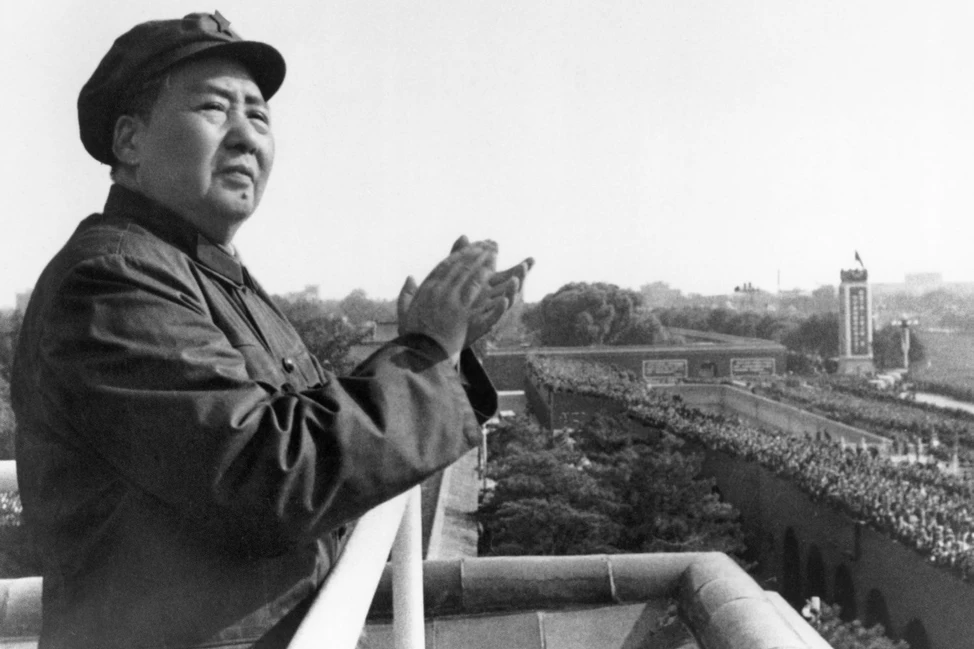 Мао Цзэдун в военной форме и кепке стоит на трибуне, внизу видна многотысячная толпа. Историческое ч/б фото.