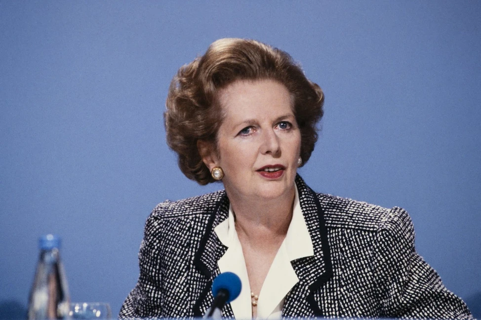 Маргарет Тэтчер выступает на пресс-конференции. Женщина в чёрно-белом пиджаке с жемчужными серьгами у синего микрофона.