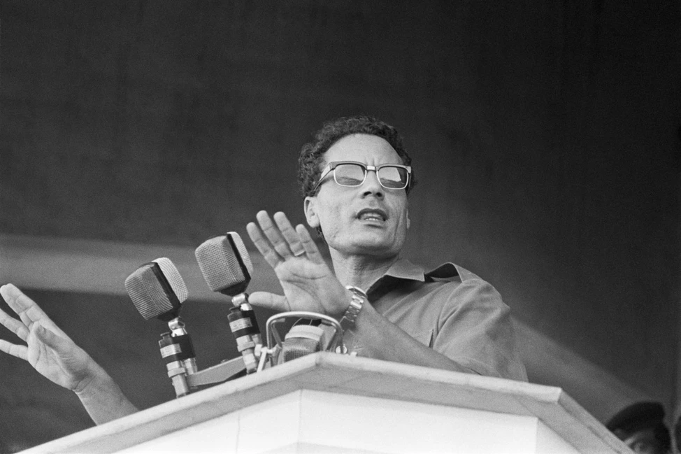 Муаммар Каддафи в тёмных очках выступает с речью за трибуной с микрофонами, 1973
