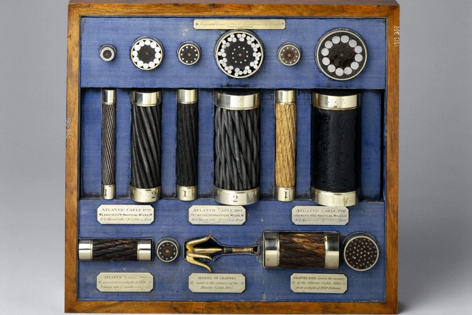 Образцы трансатлантических телеграфных кабелей (1858–1866): срезы, сегменты и модель грапнеля в витрине на синем фоне.