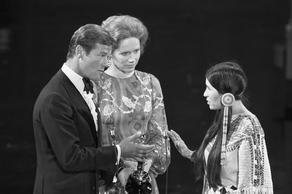 Черно-белое фото: Роджер Мур держит статуэтку Оскар, рядом Лив Ульман. Напротив стоит Сашин Литтлфезер в традиционном наряде.