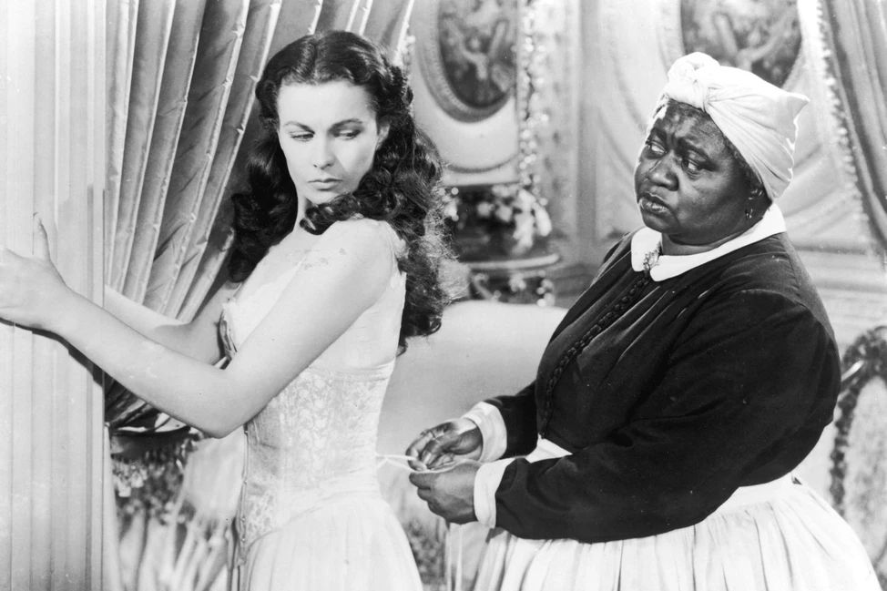 Кадр из классического фильма: женщина в элегантном платье у шторы и горничная в тёмной форме с белым головным убором в роскошном интерьере.