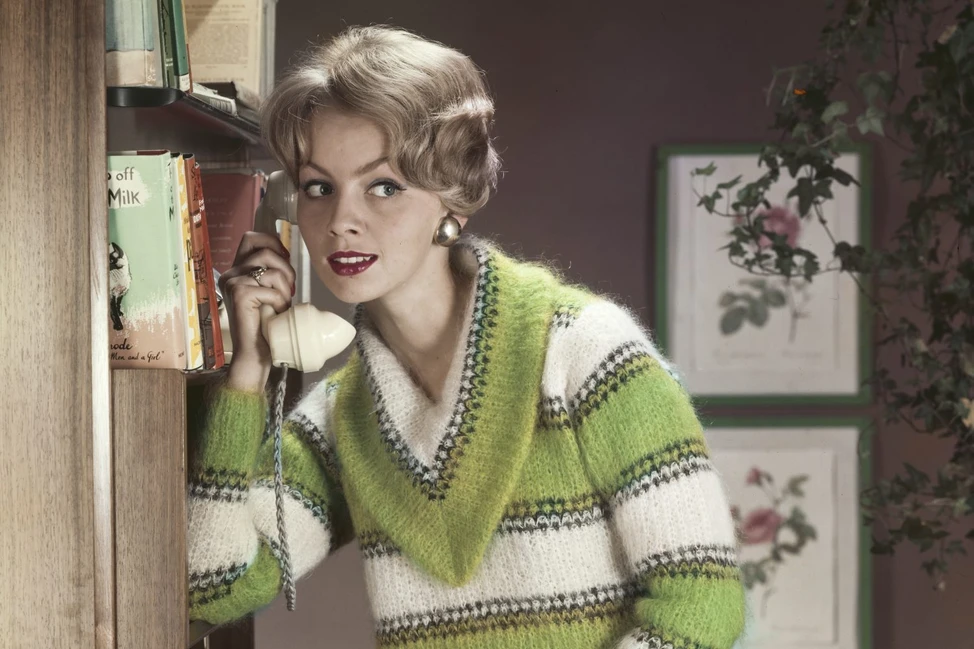 Молодая женщина в полосатом зелёно-белом свитере разговаривает по винтажному телефону у книжной полки