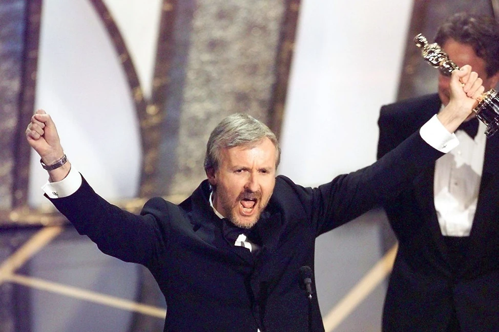 Режиссёр с седыми волосами и бородой в смокинге триумфально поднимает руки вверх, держа статуэтку Оскар, выражая радость победы.