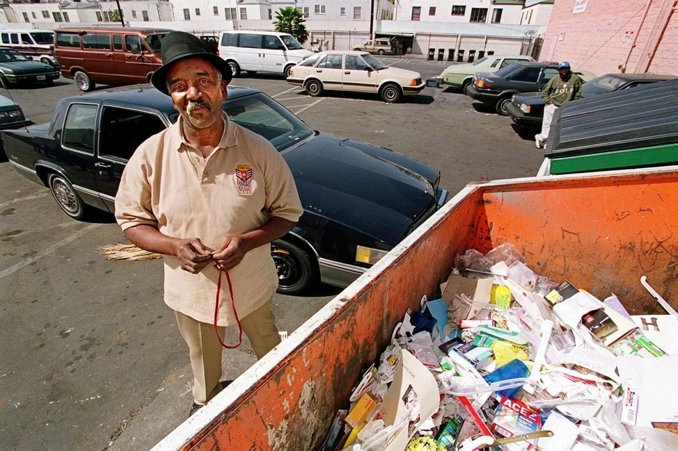 Мужчина в шляпе стоит рядом с оранжевым мусорным контейнером, заполненным отходами, на парковке с автомобилями на заднем плане.