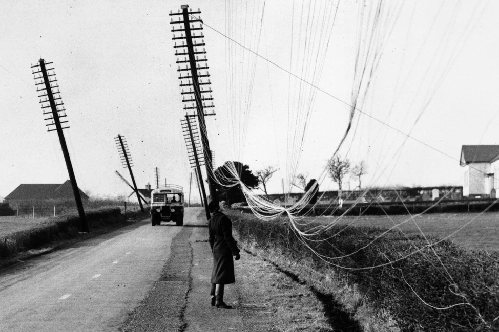 Женщина в тёмном пальто у обочины, накренившиеся телеграфные столбы с запутанными проводами, на дороге автомобиль