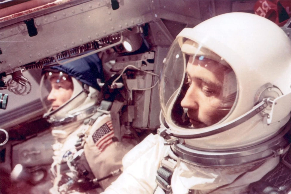 Два астронавта в белых скафандрах с шлемами внутри космической капсулы, на форме американский флаг