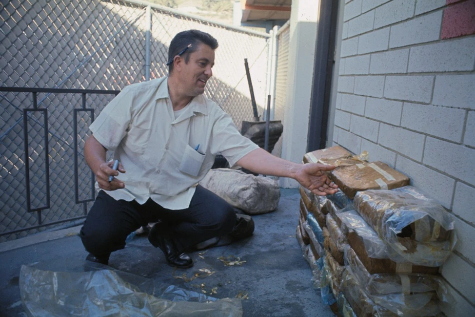 Сотрудник в светлой рубашке осматривает перевязанные свёртки у кирпичной стены и сетчатого забора, 1969