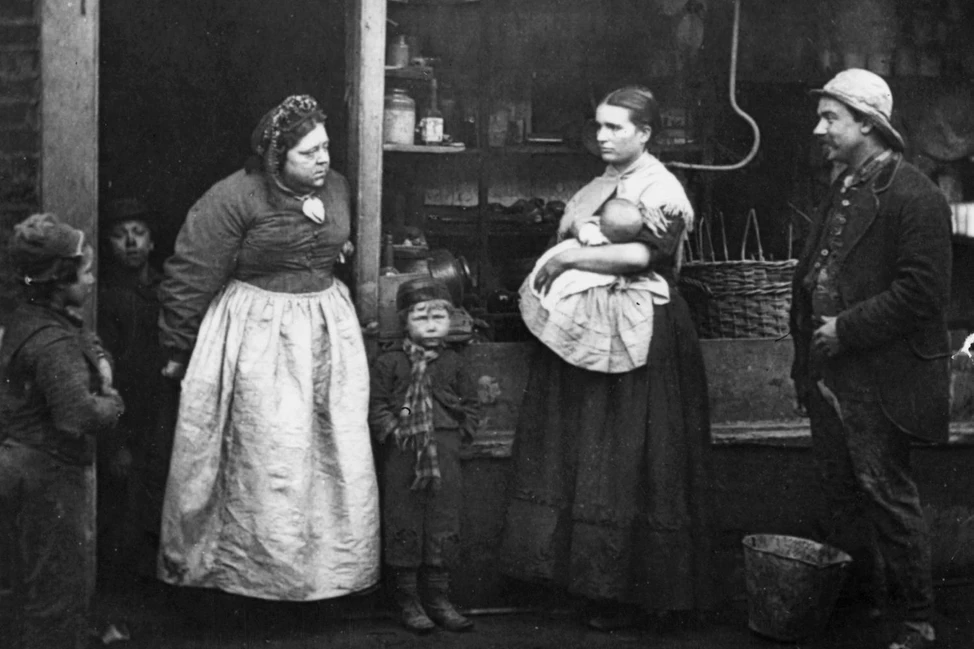 Люди викторианской эпохи у лавки: женщины в фартуках с детьми, мужчина в рабочей одежде, 1870-е годы