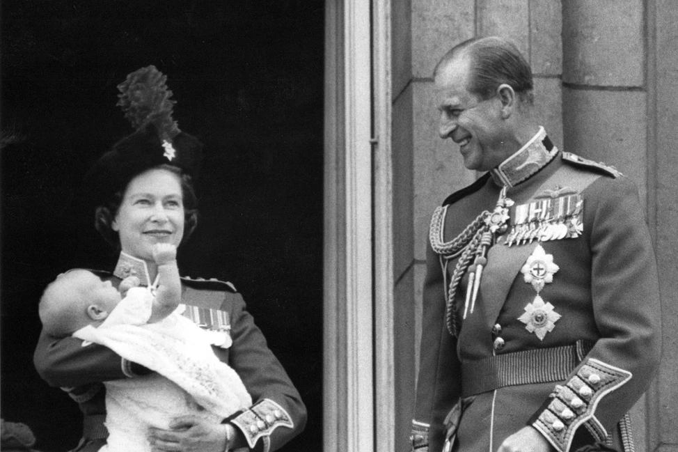 Елизавета II с младенцем на руках стоит рядом с принцем Филиппом в парадной форме у дворцового входа