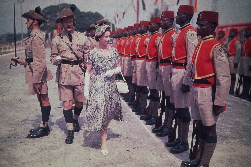 Елизавета II проходит вдоль строя африканских военнослужащих в парадной форме, рядом офицеры в шортах