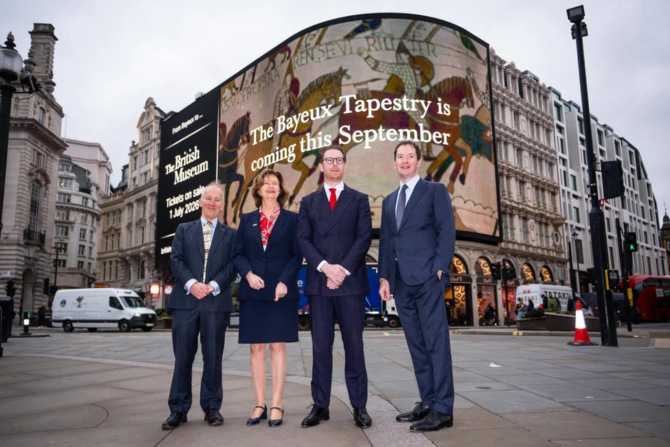 Четверо участников на Пикадилли-сёркус у экрана с анонсом выставки гобелена из Байё в Лондоне