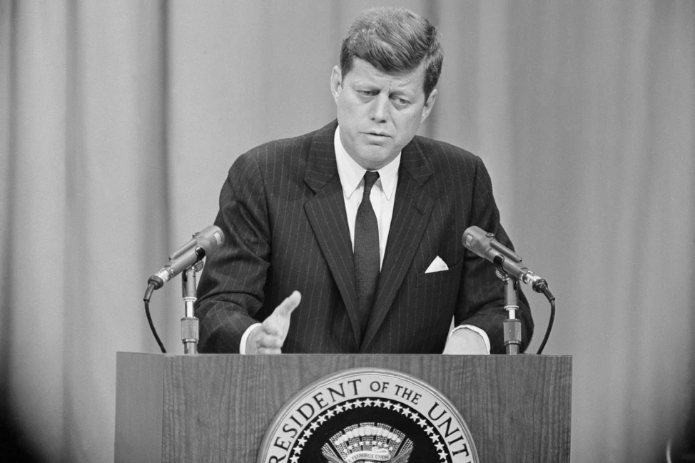 Джон Кеннеди в тёмном костюме у президентской трибуны с гербом США и микрофонами, 1961 год