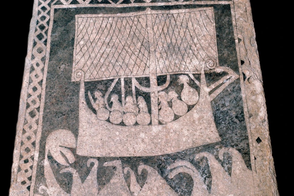 Рунический камень с резным изображением драккара под парусом и фигурами людей, орнаментальная рамка по краям