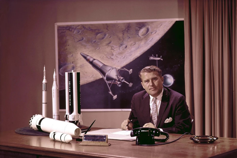Вернер фон Браун за столом с моделями ракет, на стене плакат с Луной и космическим кораблём