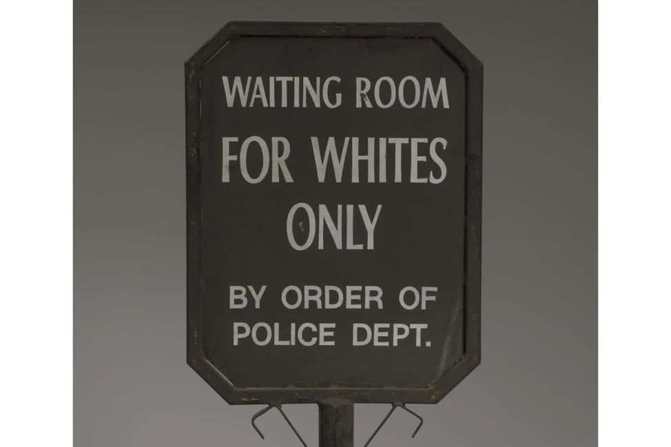 Восьмиугольная тёмная вывеска с белым текстом WAITING ROOM FOR WHITES ONLY BY ORDER OF POLICE DEPT