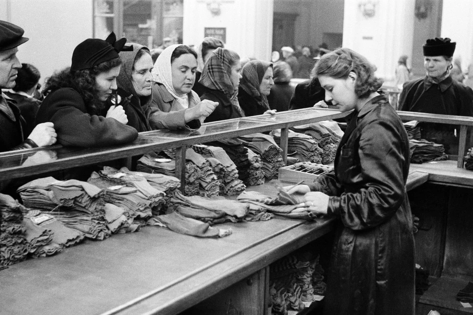 Покупательницы у прилавка с текстильными товарами в советском магазине, продавщица обслуживает клиентов, середина 1950-х